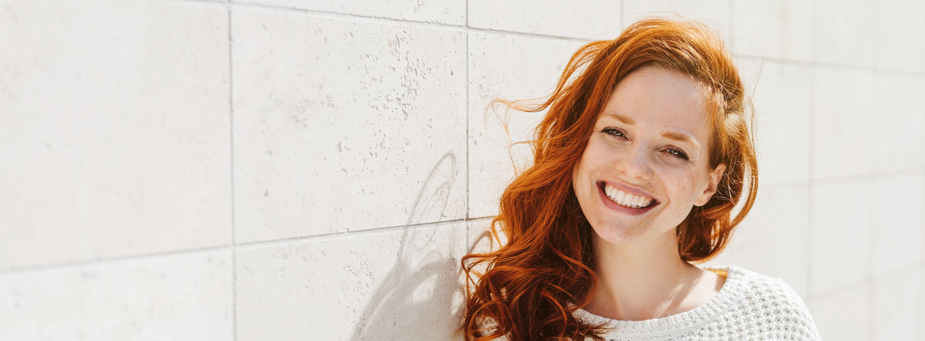 The image is a photograph of a woman with light skin, smiling at the camera. She appears to be in her late twenties or early thirties and has long hair. Her eyes are looking directly at the camera, and she is holding up her index finger near her mouth as if she's making a point or emphasizing something. The background is plain and light-colored, which suggests that this could be a stock photo used for various purposes such as advertising, personal branding, or lifestyle content.